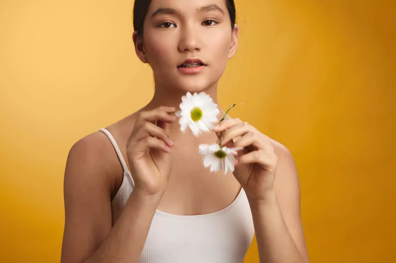 Asian woman holds white daisies against a warm orange background, creating a calm, fresh mood with graceful pose and soft lighting, highlighting natural beauty and subtle emotion in a minimalist