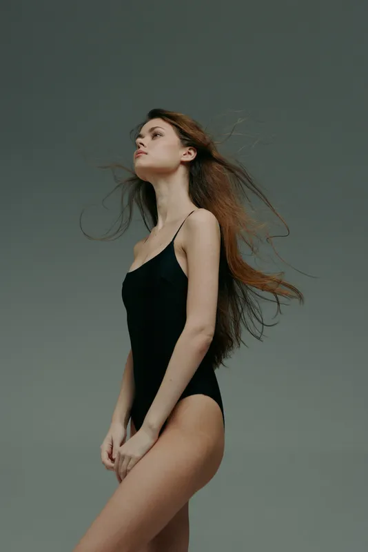 Young woman in black bodysuit posing with flowing hair against gray background, elegant and confident posture.