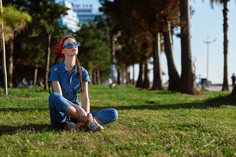 Lifestyle woman in modern boho-western denim street style sitting on grass outdoors wearing sunglasses and bandana, captured with film color tones in sunny park setting.
