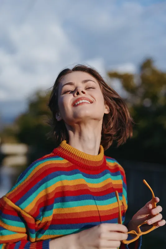Woman smiling in her mid 20s wearing a colorful striped sweater and holding sunglasses outdoors, short hair, joyful expression, casual outfit, lifestyle portrait in park.