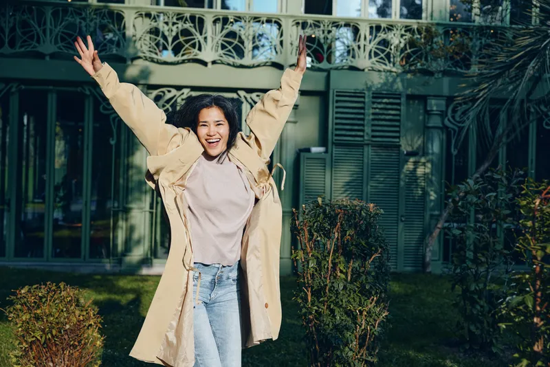 Joyful woman outdoors in a long coat with arms raised, celebrating a moment outside a modern house and garden, conveying happiness, freedom, and spontaneous enthusiasm