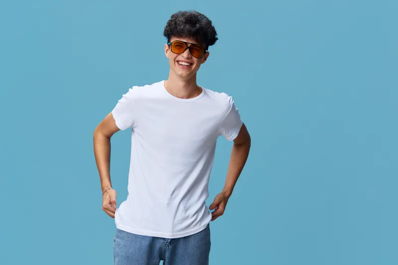 smiling man wearing sunglasses and a white t-shirt stands confidently against blue background with casual jeans, expressing happiness and youthfulness.