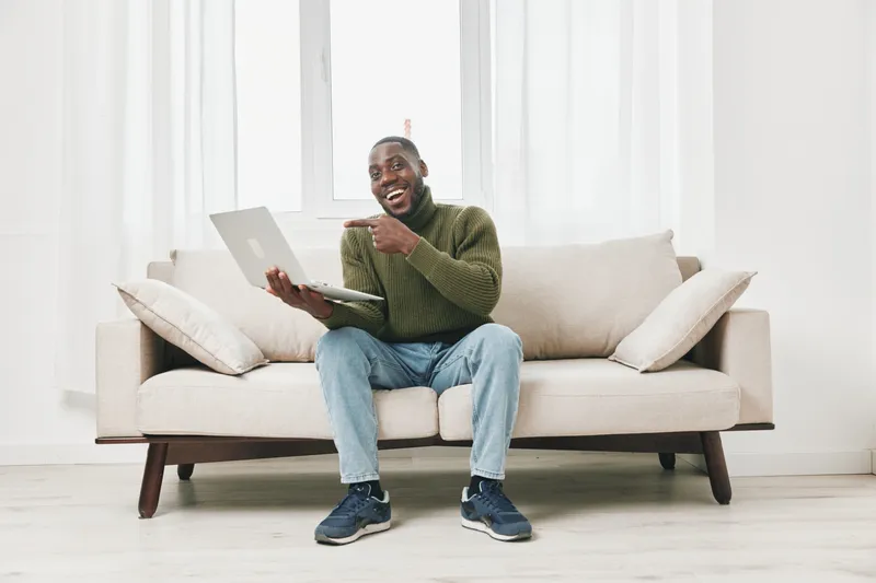 Happy young man working on a laptop at home, wearing a green sweater and jeans, sitting on a light sofa in a bright, minimalist living room