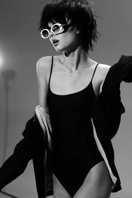 Black and white portrait of a young woman wearing sunglasses and a swimsuit with a jacket draped over her arm.
