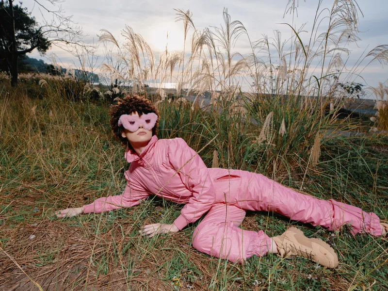 A masked figure wearing a pink costume lies on grassy ground in an outdoor scene, playful yet dramatic, with tall reeds around, creating an artistic portrait full of surreal fashion and nature