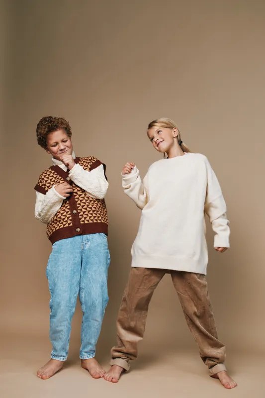 Two children pose in cozy winter outfits against a neutral backdrop, one in a patterned knit vest and jeans, the other in an oversized cream sweater and warm trousers.