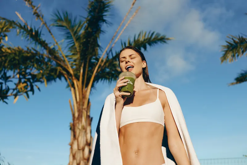 Active outdoor lifestyle moment with a young woman in a white bikini top, towel over shoulders, drinking a drink of green smoothie under sunshine, skies with a backdrop of palm trees and blue vacation