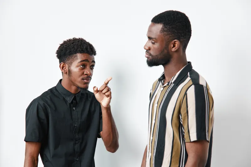 Two African American men with dark skin having a serious conversation in casual clothing against white background. One man gestures while the other listens attentively.