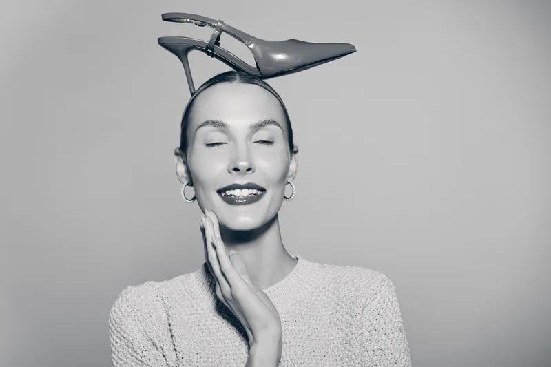 Young white woman with fair skin and closed eyes smiles gently while balancing a high heel shoe on her head in a minimalist studio. Concept for fashion, beauty, and creativity.