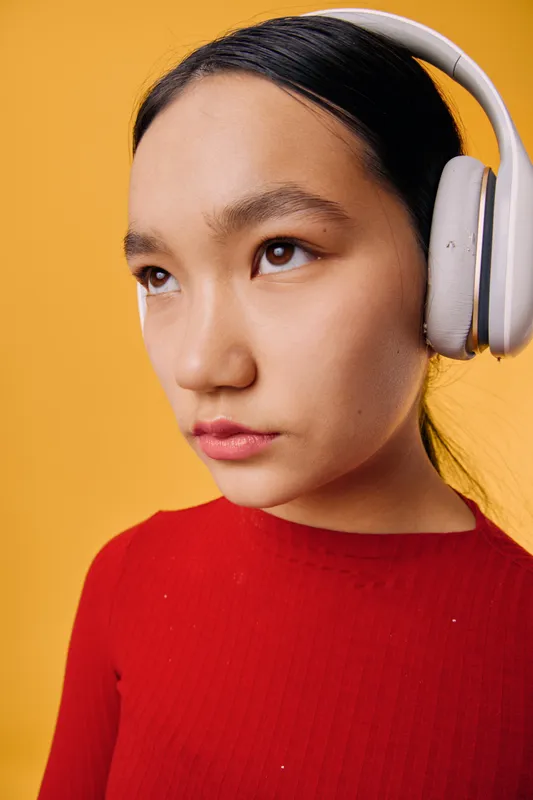 Young woman wearing wireless headphones in red sweater looking sideways with focused expression on yellow background. Closeup portrait of female listening to music with modern audio device.