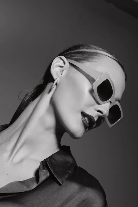 Elegant Caucasian woman with pale skin and slicked-back hair wearing sunglasses and dark satin shirt in black and white studio portrait.