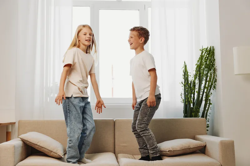 Happy children jumping on sofa in bright living room with natural light, casual clothes and playful mood at home interior.