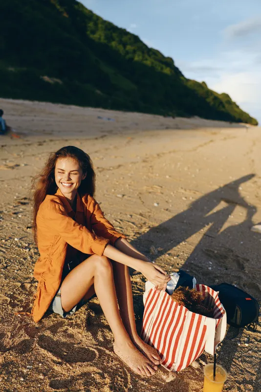 Smiling woman sitting on beach sand in sunshine, holding striped bag, relaxed outdoors, natural mood, warm light, gentle breeze, distant green hills, soft shadows, sunlit summer scene
