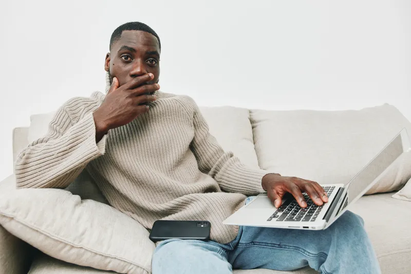A surprised man sitting on a couch with a laptop, covering his mouth in disbelief casual outfit, modern decor, neutral colors