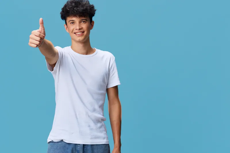 smiling young man in casual white t-shirt giving thumbs up gesture with blue background, positive expression and curly hair, youthful and cheerful mood.