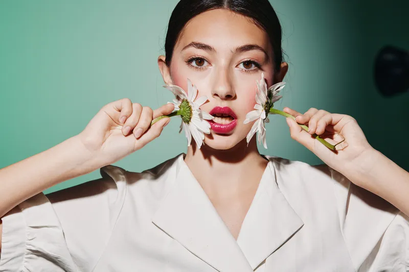Young woman in creative fashion posing with two white daisy flowers by her cheeks on green background. Concept of beauty, style, floral accessories, and innovative portrait photography.