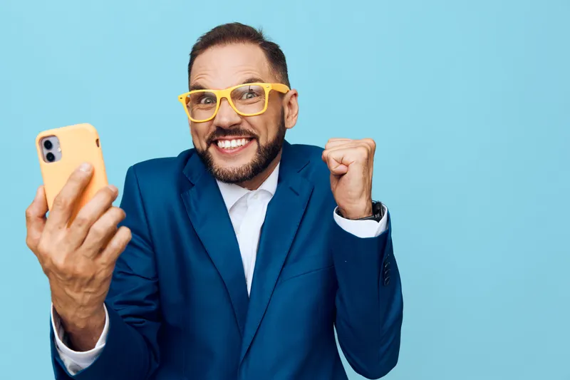 A confident business professional man stands against a solid blue background, showcasing excitement with a bright smile, raised fist, and energetic gesture.