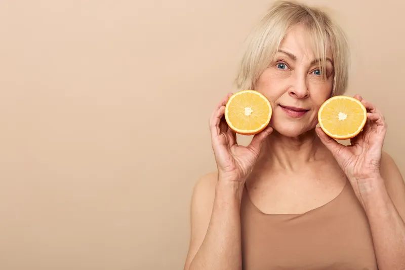 Mature woman with blonde hair holds two halves of fresh orange near face smiling gently on beige background. Concept of healthy lifestyle, natural beauty, and vitamin C benefits.