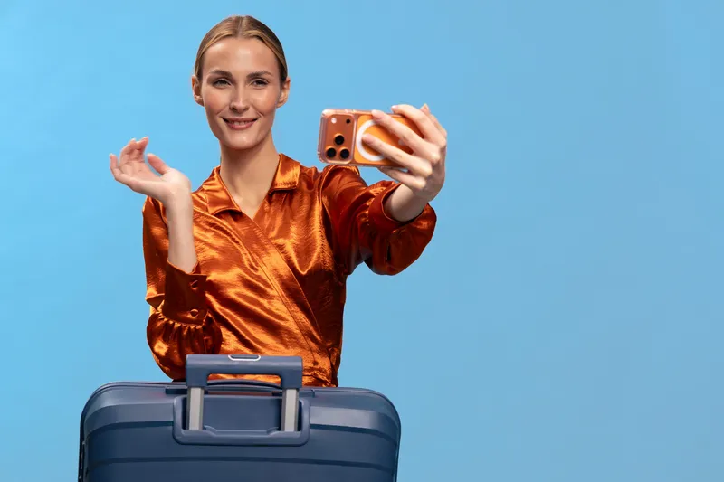 Young woman in shiny orange blouse takes selfie with smartphone while standing behind blue suitcase on blue background. Concept of travel, vacation, tourism, and modern lifestyle.