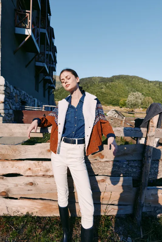 Woman leans on wooden fence in countryside with denim jacket, boots and white pants, posing relaxed against mountains and sunlight for fashion portrait, rural lifestyle and travel.