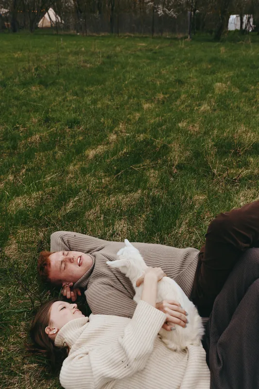 Young couple lies on grass in cozy sweaters holding a white baby goat enjoying a peaceful lifestyle moment outdoors in early spring with relaxed and happy expressions.