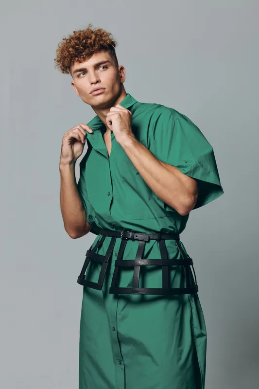 Man with curly hair and serious face posing in green shirt dress with short sleeves and belt in geometric design on gray studio background posing confidently and fashionably.