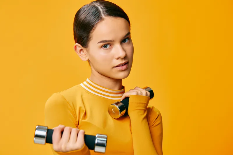 Female fitness enthusiast lifts dumbbells in a bright orange studio, displaying focus during a strength training routine, showcasing balanced posture, form, and determined workout momentum.