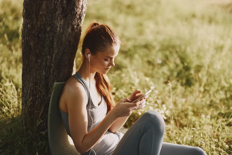 A young woman enjoying a sunny day outdoors, sitting against a tree while using her smartphone, embodying a relaxed and healthy lifestyle