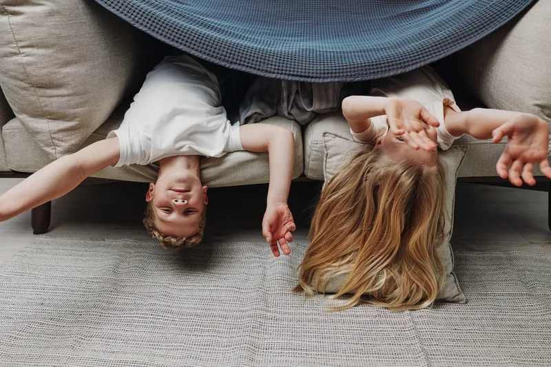 Two children with blonde hair lie upside down on a sofa covered with a blue blanket, playing and reaching out with their hands in a cozy living room environment.