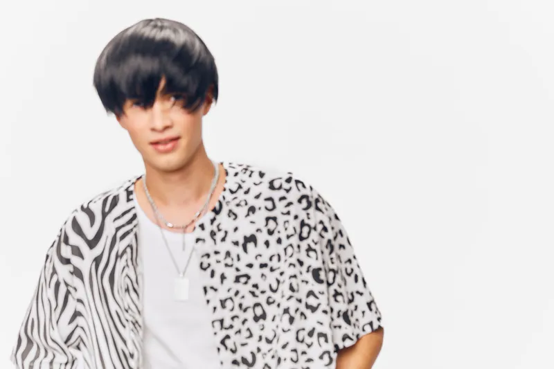 young man wearing patterned animal print shirt with layered necklaces posing against white background, trendy hairstyle and casual style