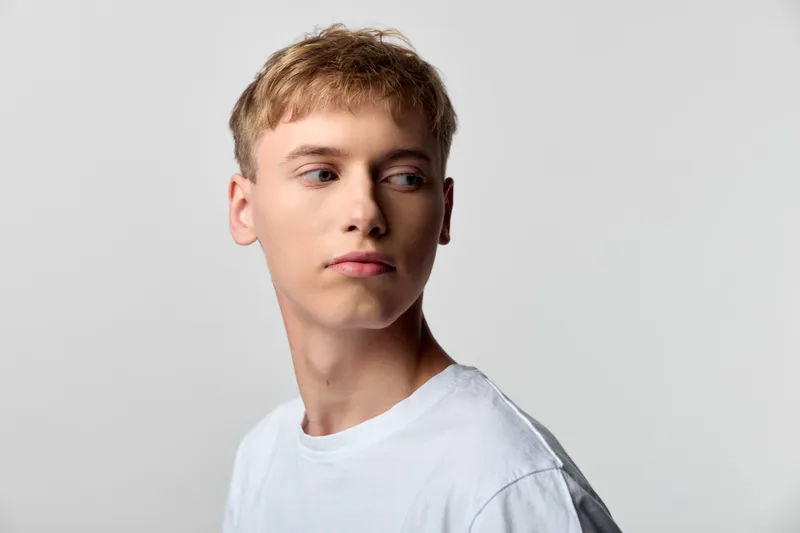 Man with short blond hair wearing a white shirt looks away against a neutral studio backdrop, conveying quiet contemplation, clean minimal style, and versatile aesthetic for stock use