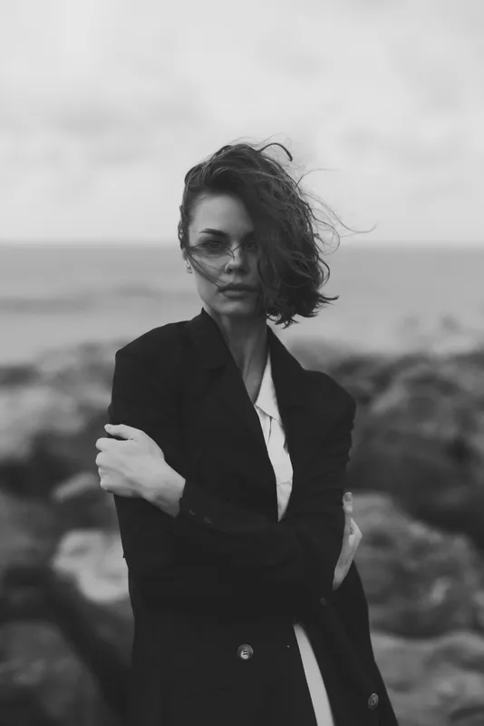 Monochrome image of a woman in a tailored coat standing on rugged rocks by the sea, wind swept hair, confident pose, moody coastal fashion photography