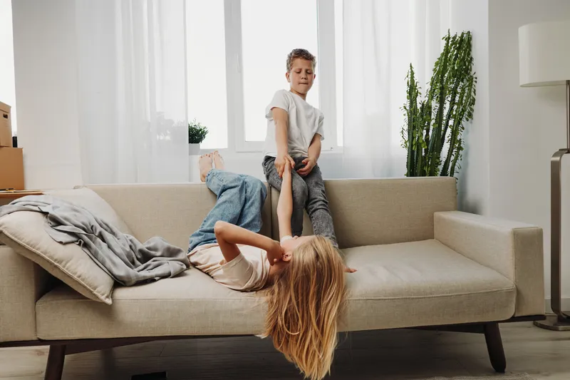 Playful children in casual clothes having fun on beige sofa in bright living room. Boy and girl enjoy playful moment together with natural light and cozy home atmosphere.
