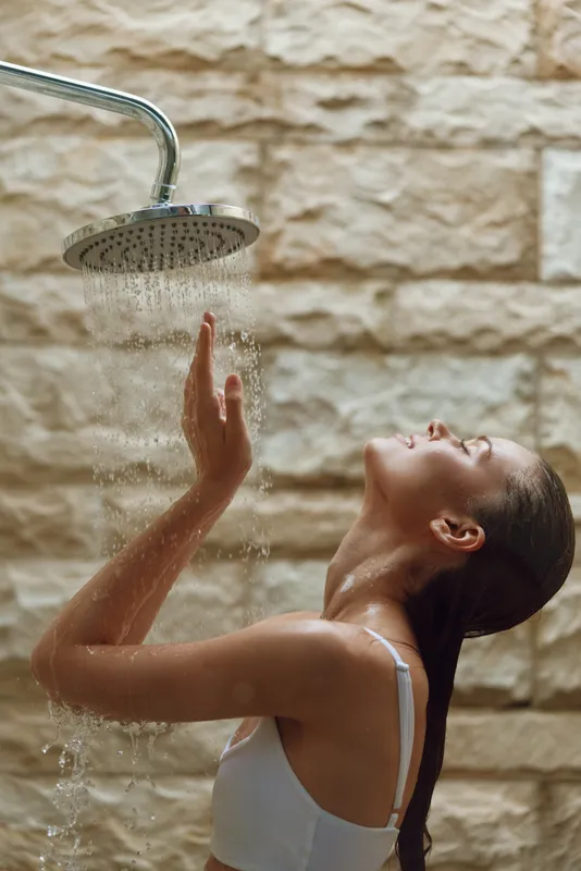Woman enjoying warm shower under rain-style showerhead, with wet hair and relaxed expression against a textured stone wall background, wellness and self-care moment.