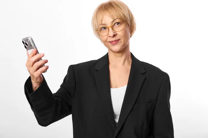 Senior woman with glasses in business blazer holds smartphone up for selfie, confident portrait, using technology communication on clean white background, isolated studio light