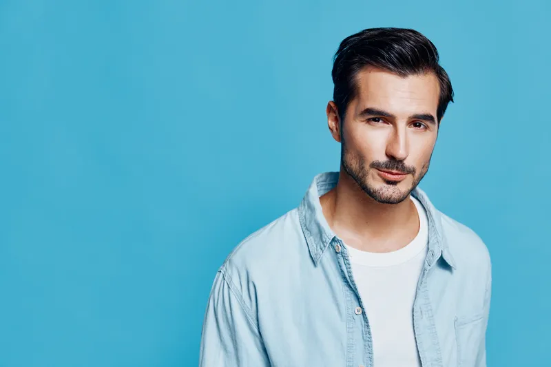 Charming young man dressed in light blue shirt poses confidently against a vibrant blue background. He gazes at the camera with a natural expression, exuding charm.