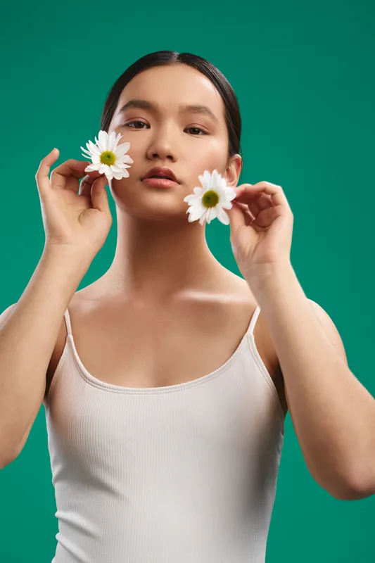 asian woman holds daisies against a vibrant teal backdrop, presenting a serene yet playful portrait. she gazes softly at the camera, creating a fresh, natural vibe of calm beauty.