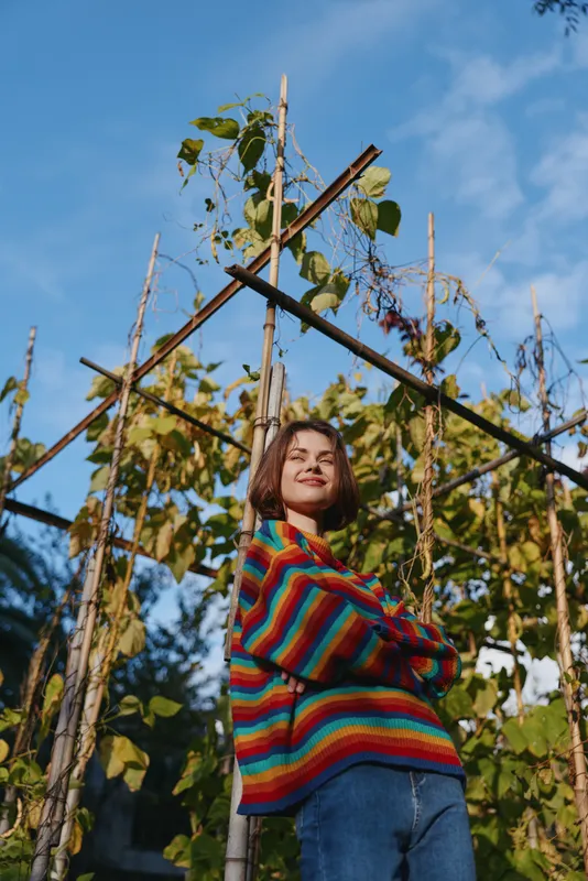 Young woman in a colorful striped sweater and jeans smiling confidently in a backyard garden by a wooden trellis, leaning with crossed arms in a sunny lifestyle portrait.