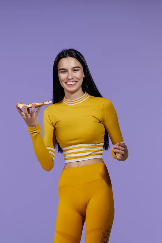 Woman sport fitness wearing yellow activewear smiling while holding a slice of pizza against a violet background, showing balance between healthy lifestyle and enjoyment.