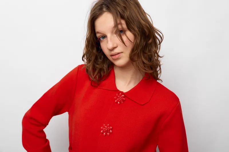 Young woman with wavy brown hair wearing a red sweater with decorative buttons poses against white background. Casual fashion portrait with natural makeup and relaxed expression.