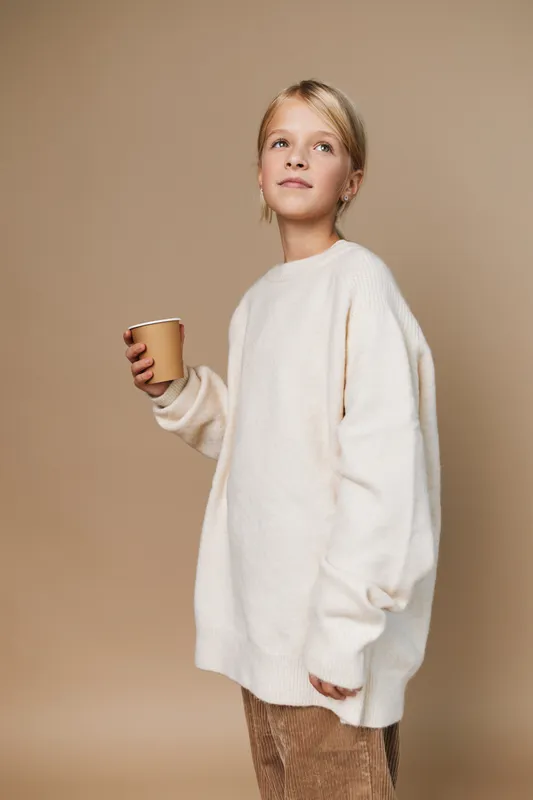 Authentic lifestyle portrait of a young girl in a cozy oversized sweater, holding a cup, conveying playful schooldays and simple everyday moments in a calm studio setting.