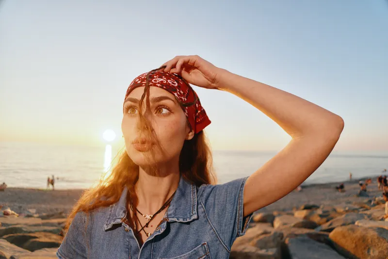 Lifestyle woman wearing modern boho-western denim and red bandana standing by the beach at sunset, film color style capturing relaxed street fashion vibes.