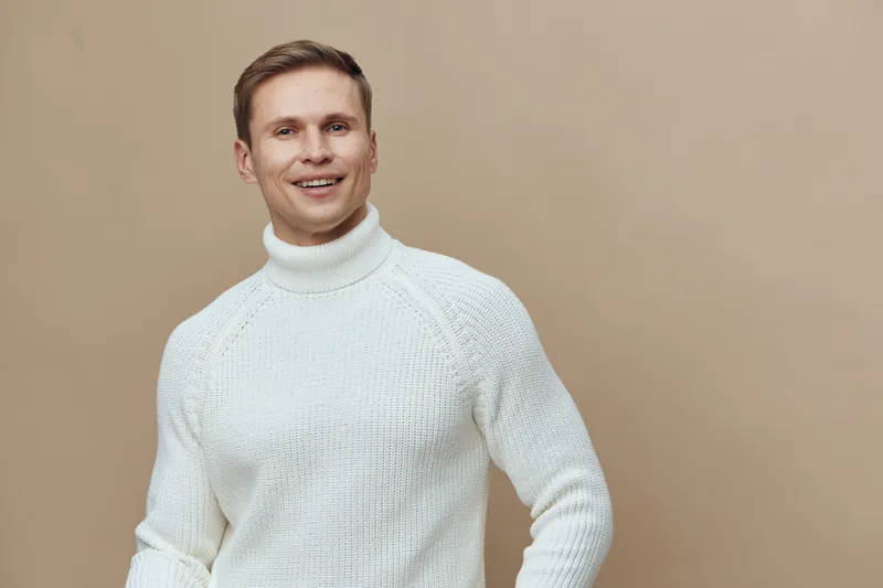 Happy smiling man wearing white knitted turtleneck sweater posing against plain beige background in studio. Casual fashion, cheerful expression, menswear, portrait shot, lifestyle concept.