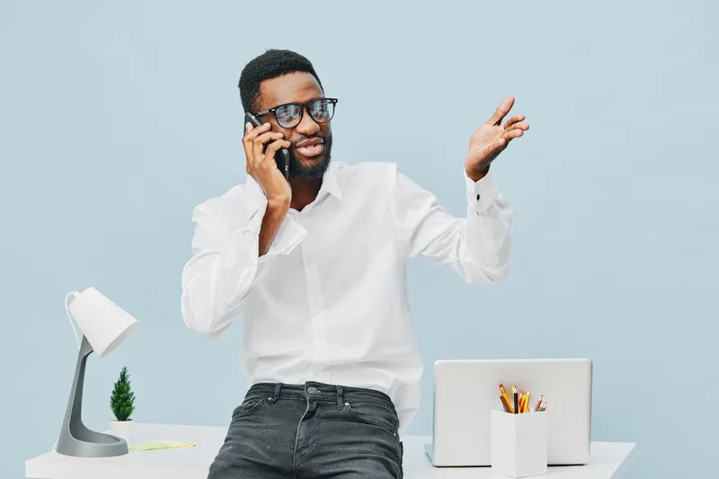 Confident young man with glasses talking on mobile phone while gesturing with his hand in a modern office space, wearing white shirt and black jeans. Business communication, professional lifestyle con
