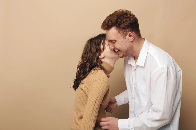 Happy young couple laughing together, woman in brown sweater whispers to man in white shirt. Intimate moment, love, friendship, and joy on beige background in casual studio setting.