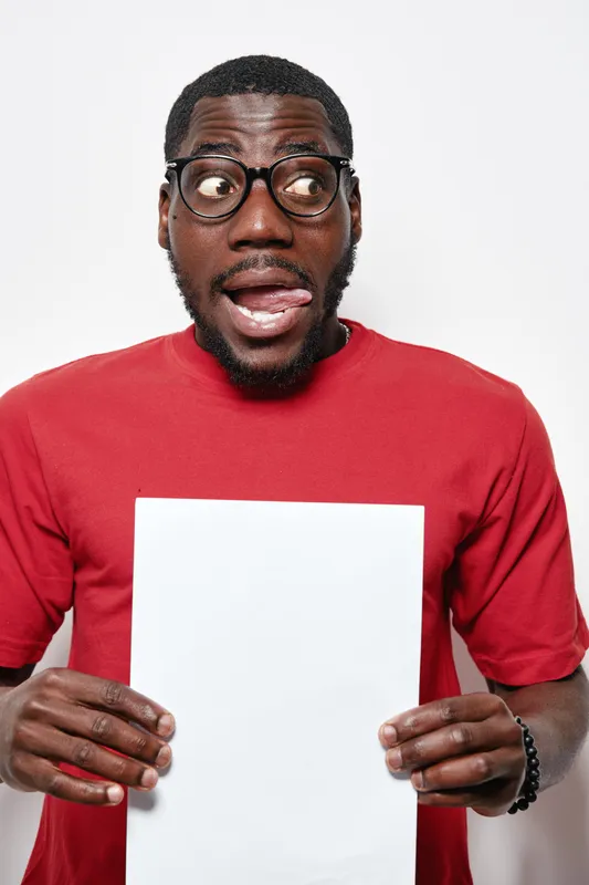 African American man with dark skin wearing glasses and red sweater holding blank white paper with playful expression on white background studio.