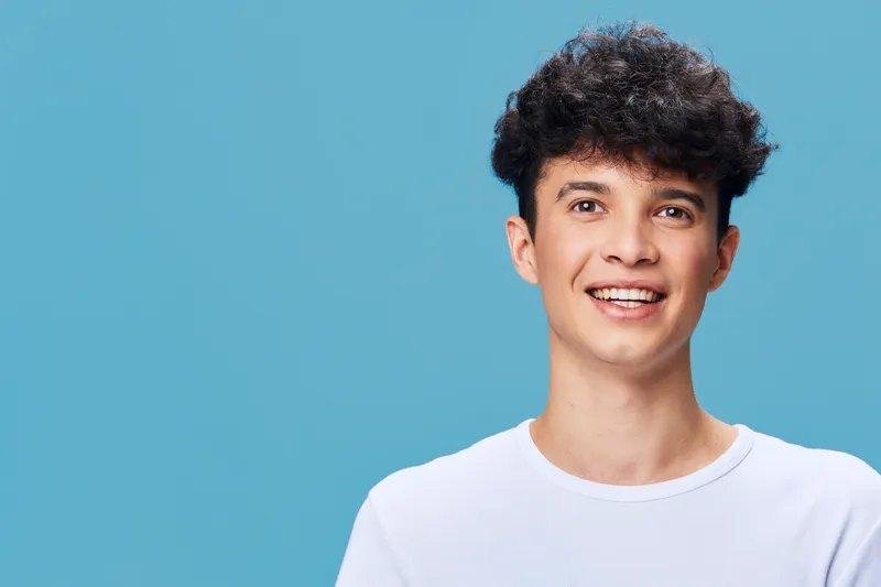 smiling young man with curly hair wearing white shirt on blue background, positive emotion, casual style, natural lighting, modern portrait