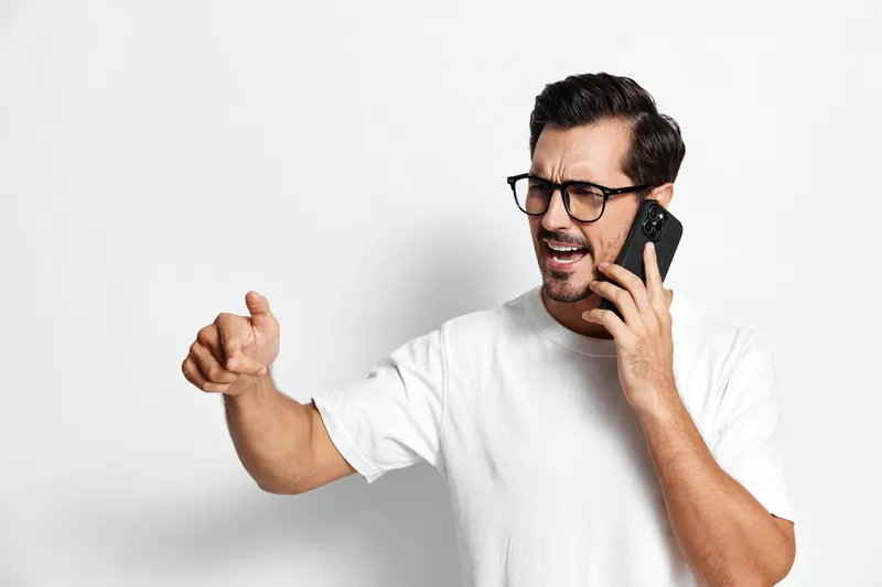 Angry young man speaking on mobile phone gesturing with hand isolated on plain solid white background. People communication concept.