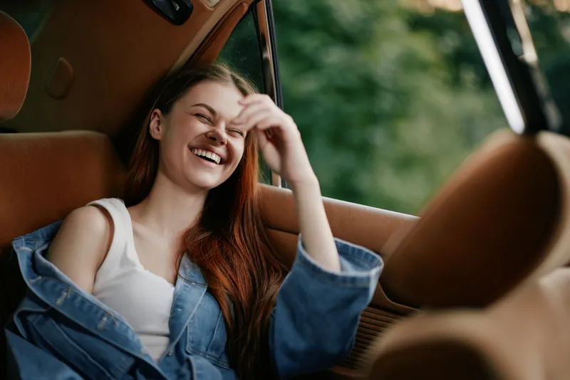 Happy young woman laughing in a car with a cozy interior, showcasing natural beauty, relaxed vibes, and outdoor greenery in a soft color palette.