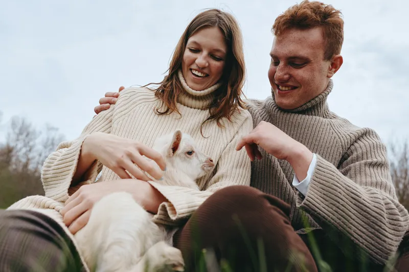Young couple in cozy sweaters happily cuddle a small white baby goat outdoors on a cloudy day, enjoying a relaxed moment in nature and connecting with the animal.